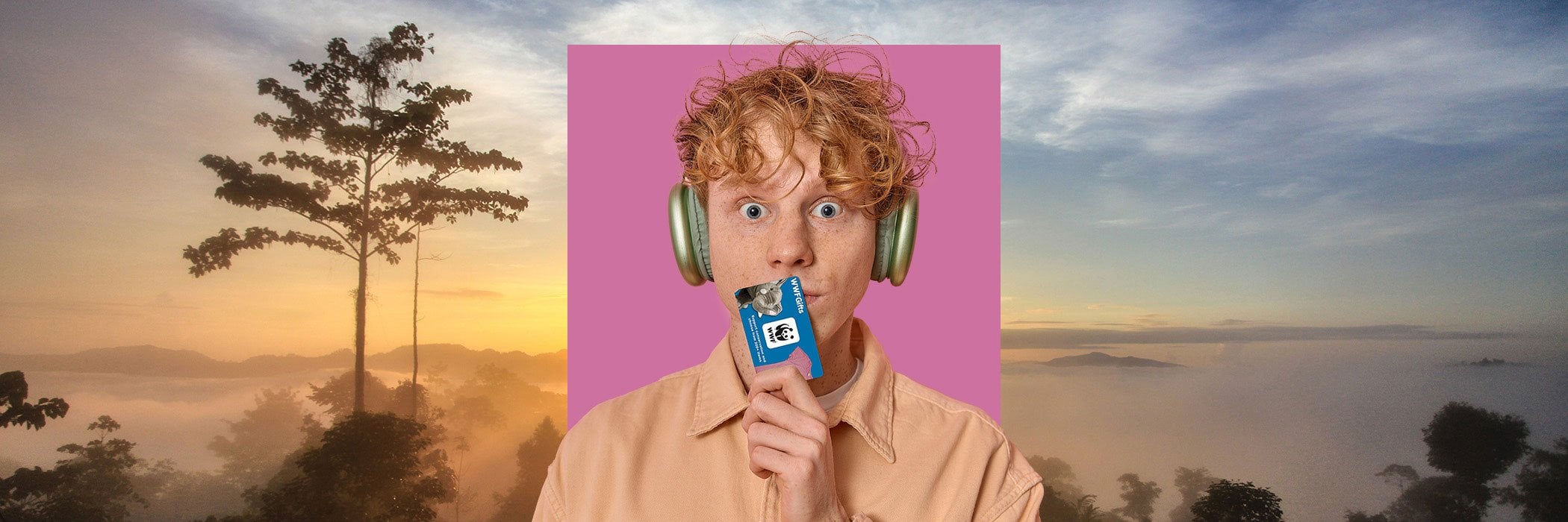 Young man with headphones on and holding WWFGifts gift card on purple square in front of epic landscape