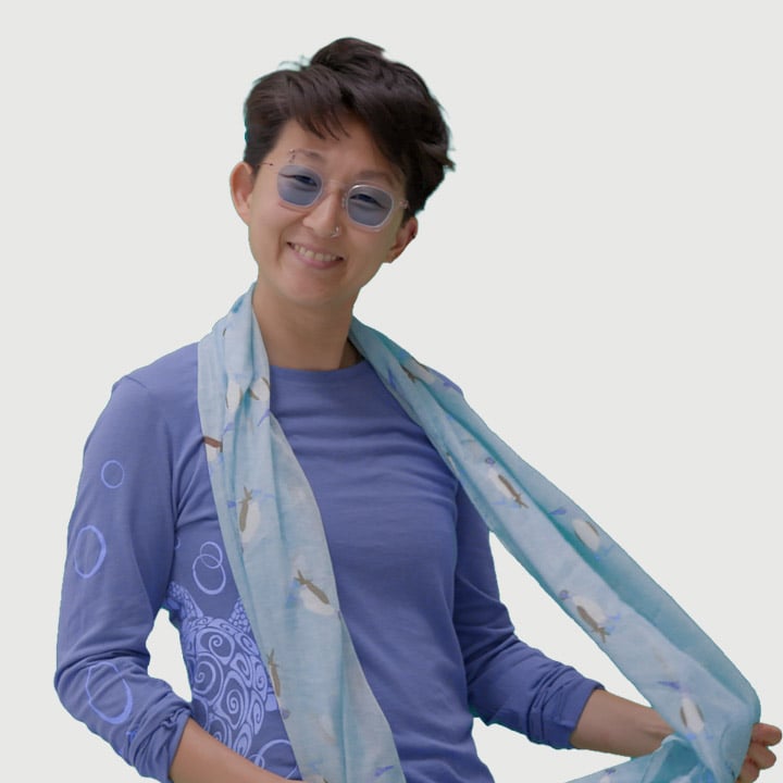 A person wearing glasses, a blue shirt with designs, and a light blue scarf smiles while looking at the camera.