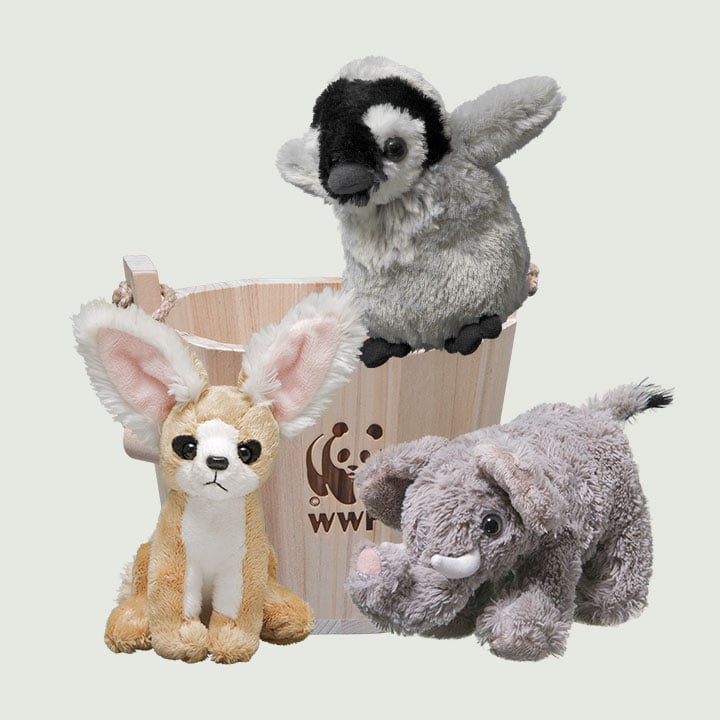 Three plush animal toys—a penguin, a fennec fox, and an elephant—are displayed with a wooden bucket featuring a WWF logo.